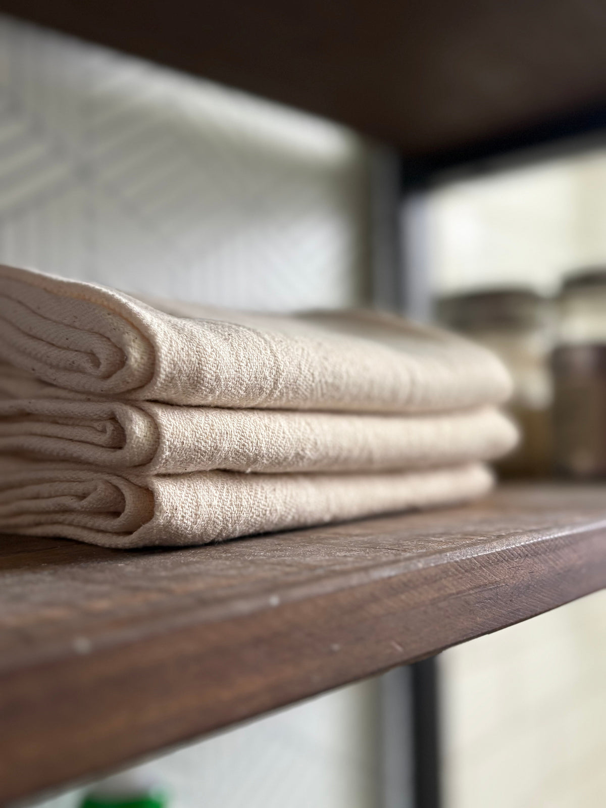 Three neatly folded beige fabric towels stacked on a dark wooden shelf, with a softly blurred indoor background.