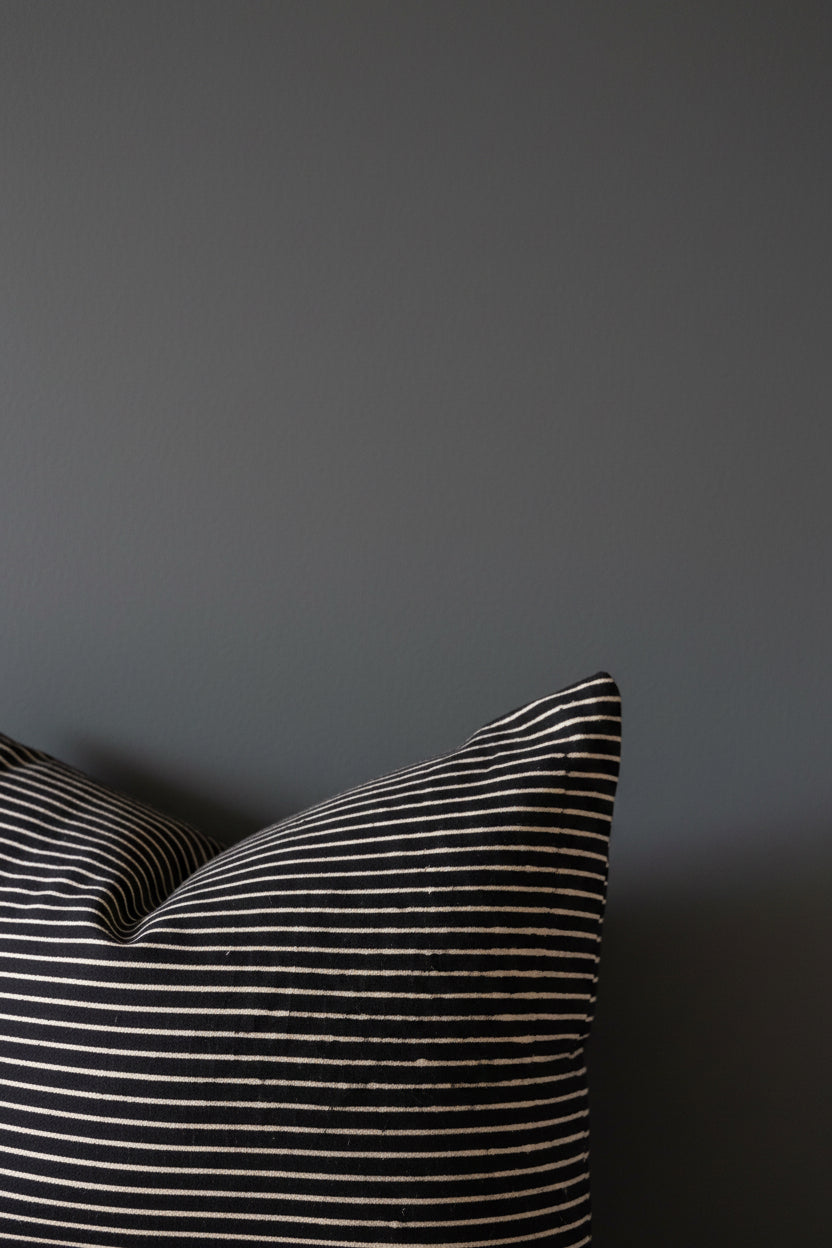 A square throw pillow with a black and beige striped pattern, resting on a wooden chair in front of a light-colored wall with framed art.