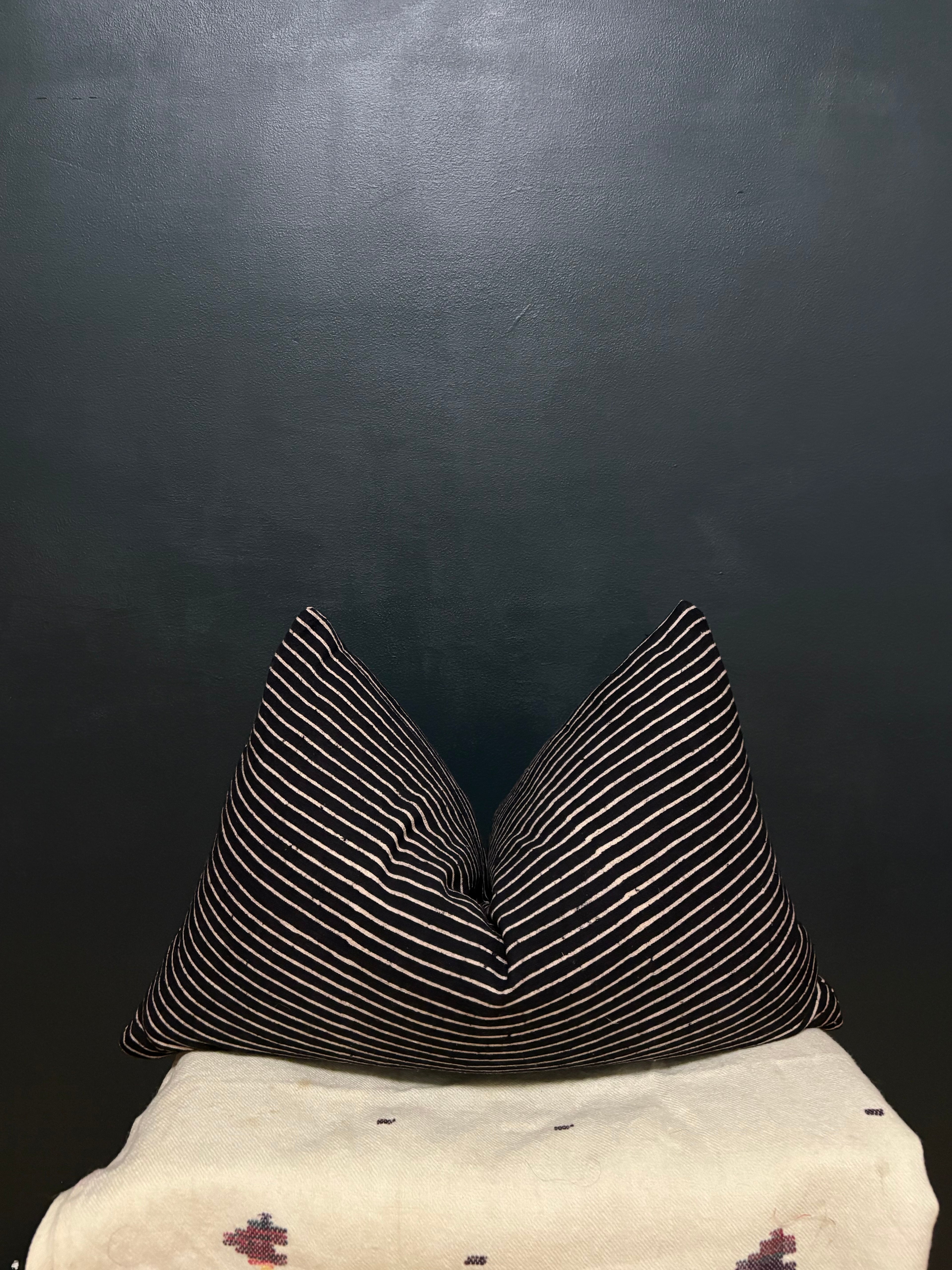 Two square throw pillows with thin white stripes on a black background, arranged on top of a cream-colored textile against a dark wall.