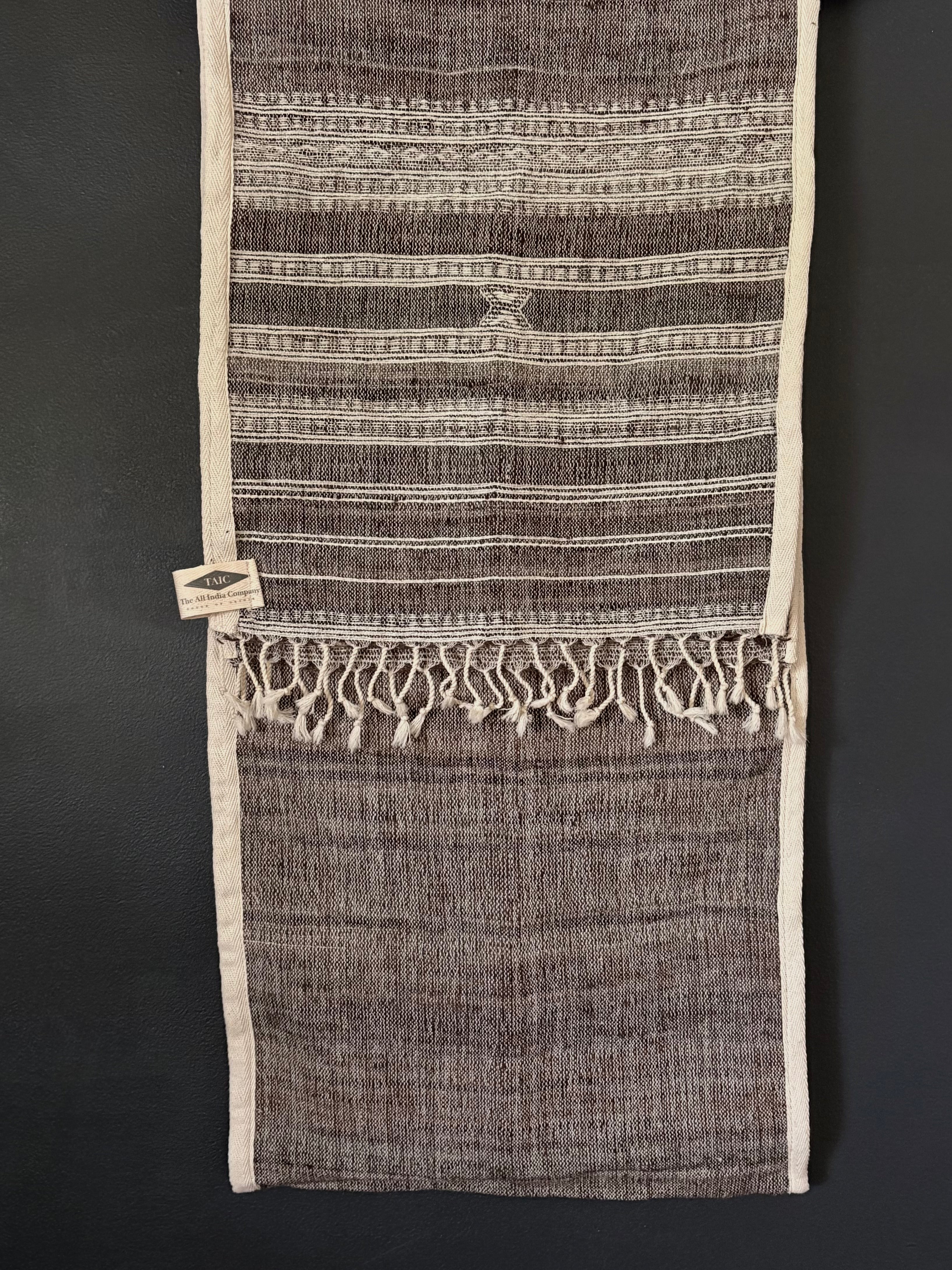 A woven textile runner with horizontal striped patterns in dark brown and cream, featuring a fringed tassel hem and a small woven label on the left side, displayed flat against a dark background.