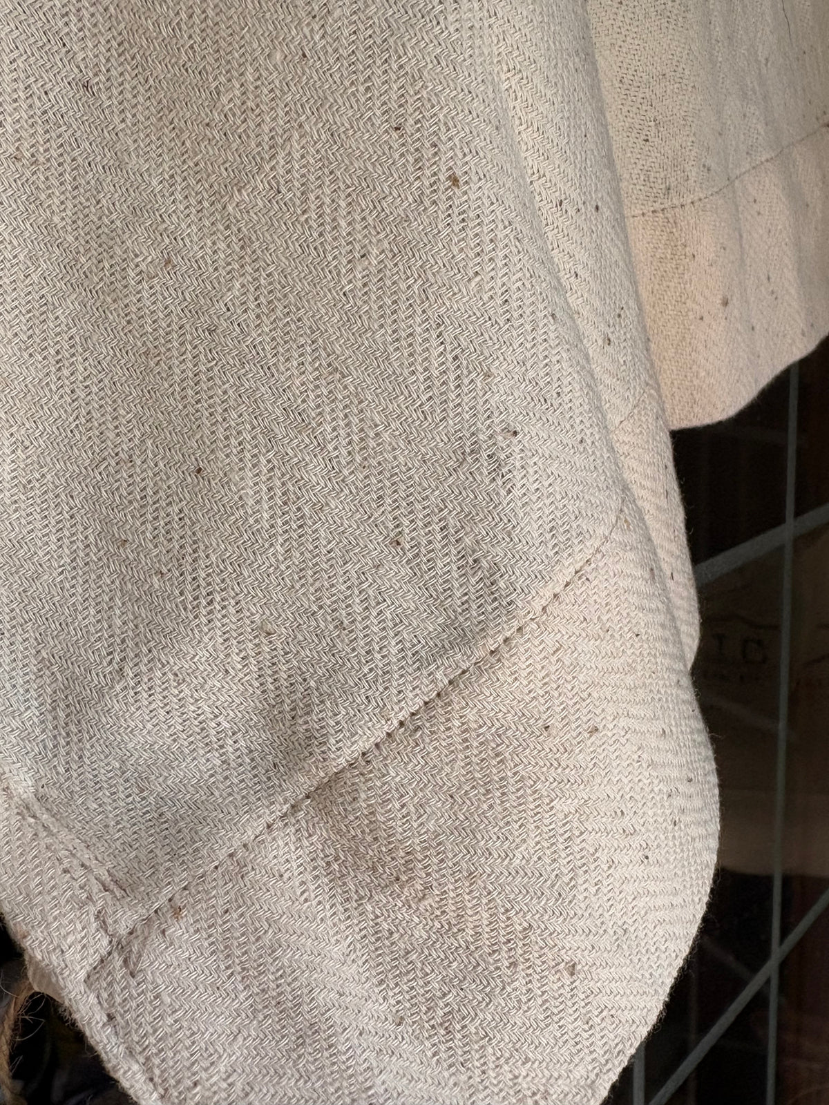 Close-up of a textured off-white woven fabric, likely linen or a similar natural fiber, showing a diagonal twill weave pattern with subtle brown specks.