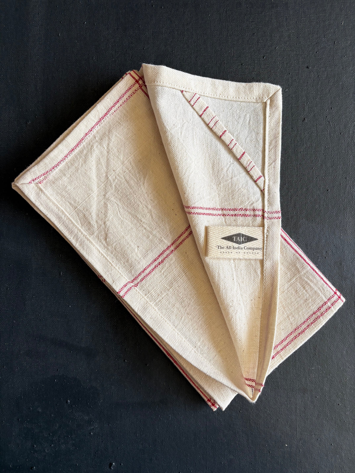 A folded cream-colored linen tea towel with red stripes and a stitched triangular detail, featuring a small paper label that reads 'The Tea Time Company', displayed on a dark textured background.