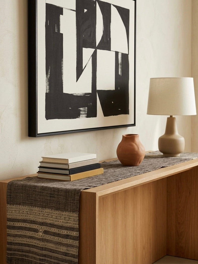 A styled interior scene showing a wooden console table with a woven runner, a stack of books, a terracotta vase, and a table lamp, beneath a large black-and-white abstract wall art print.