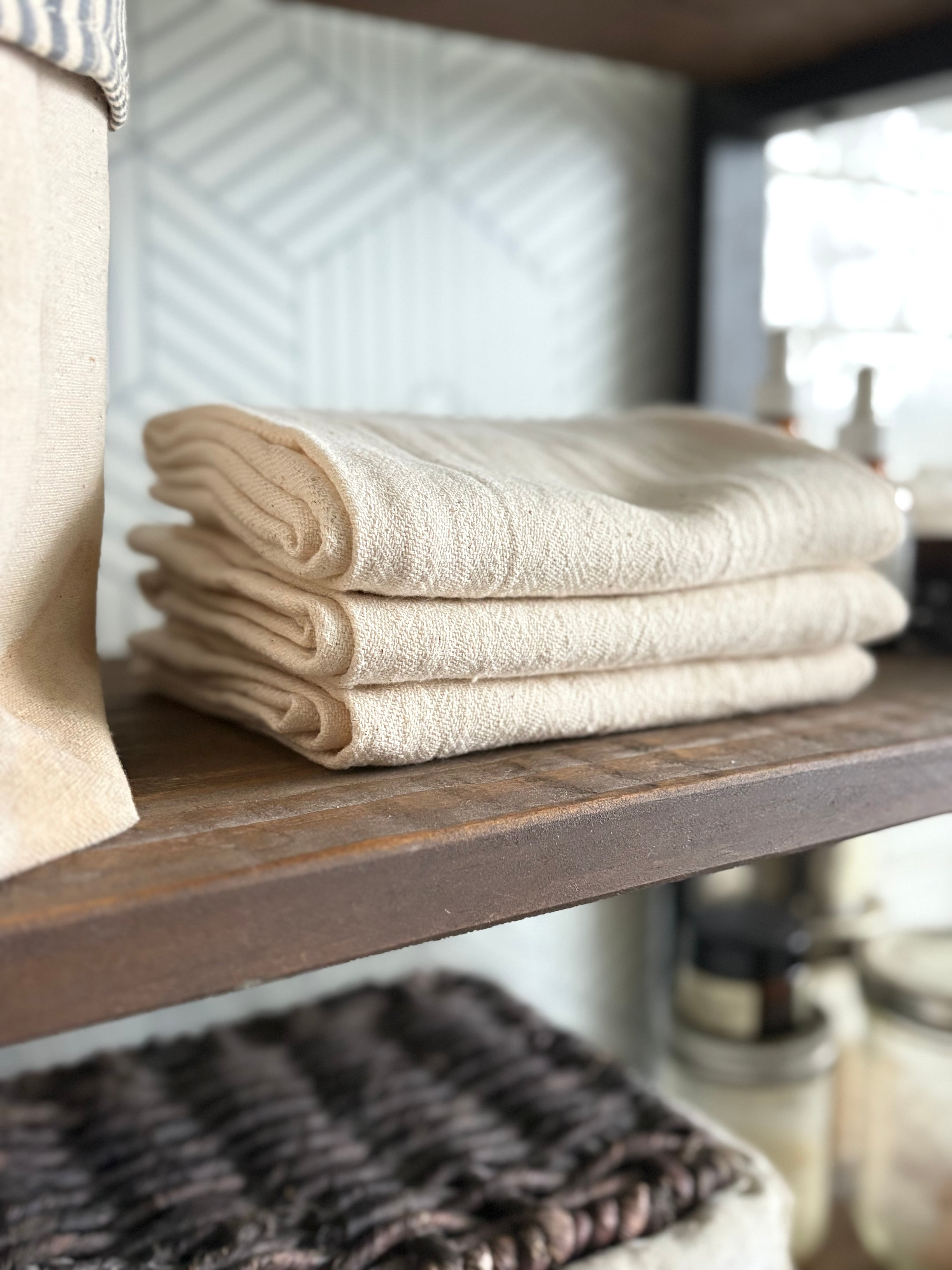 A stack of three folded cream-colored cotton towels or washcloths resting on a dark wooden shelf in a home setting.