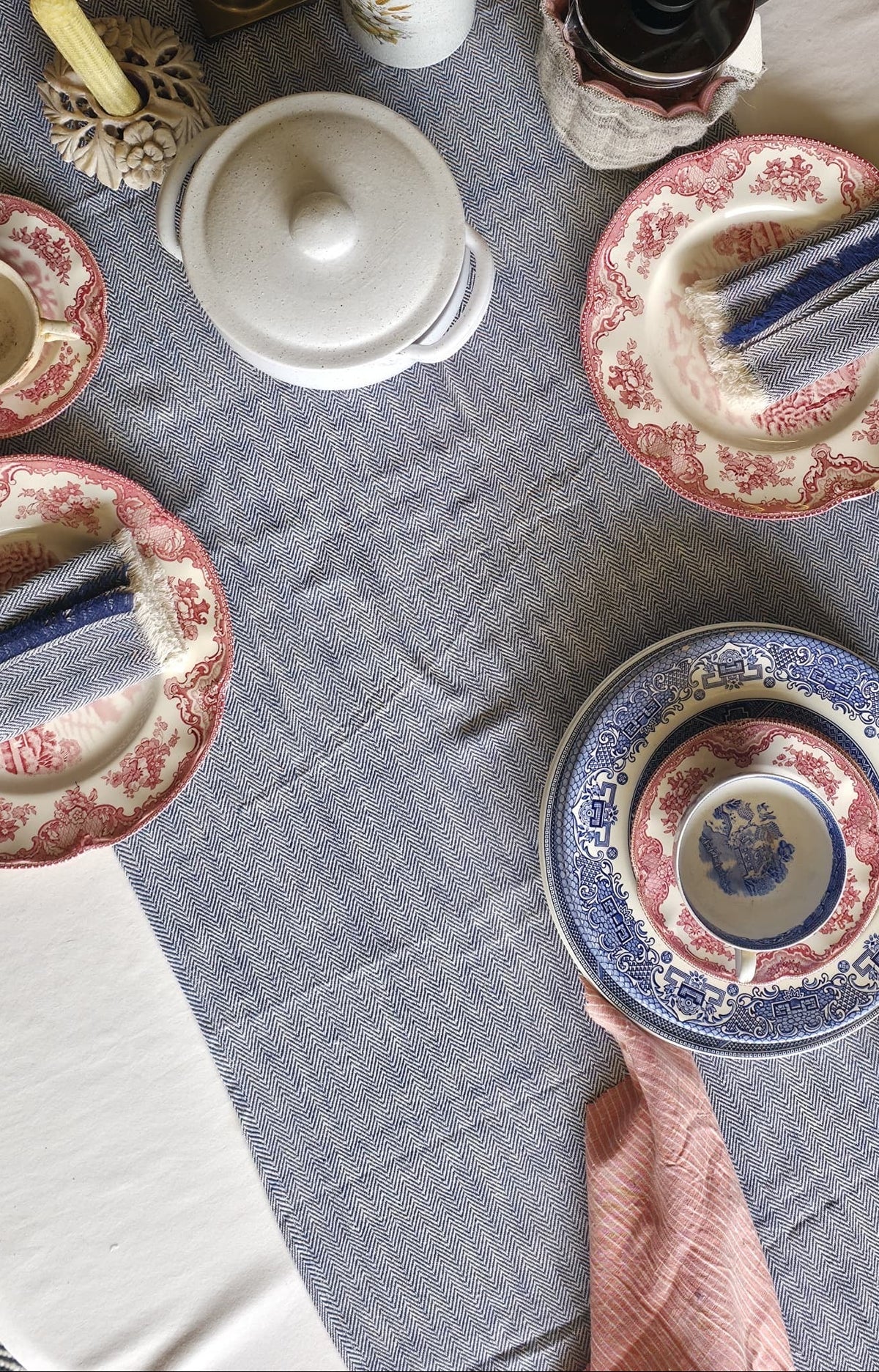 An overhead view of a dining table set with vintage-style ceramic plates, teacups, a white teapot, a candle, and a pink cloth napkin on a blue and white striped table runner.