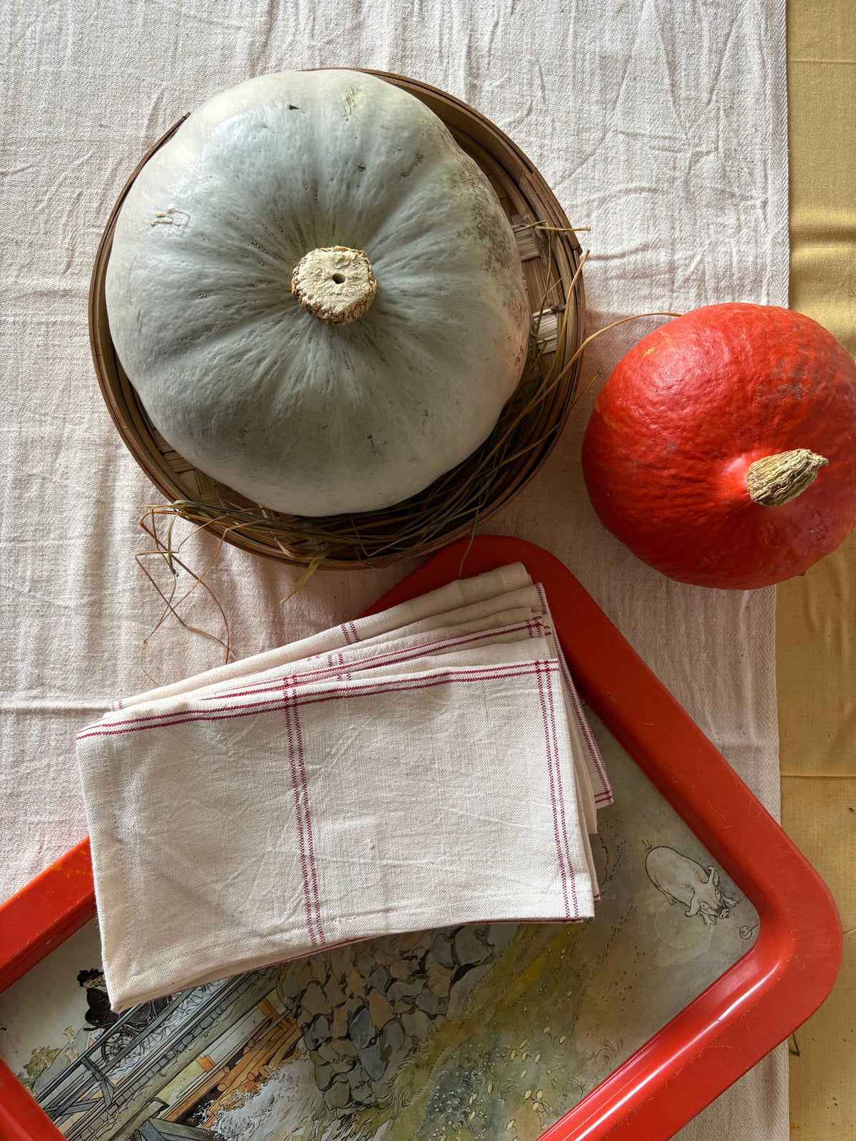 A styled flat-lay featuring a light blue-green winter squash in a woven basket, a red-orange pumpkin, and a folded cream linen napkin with an unbleached kala cotton runner.