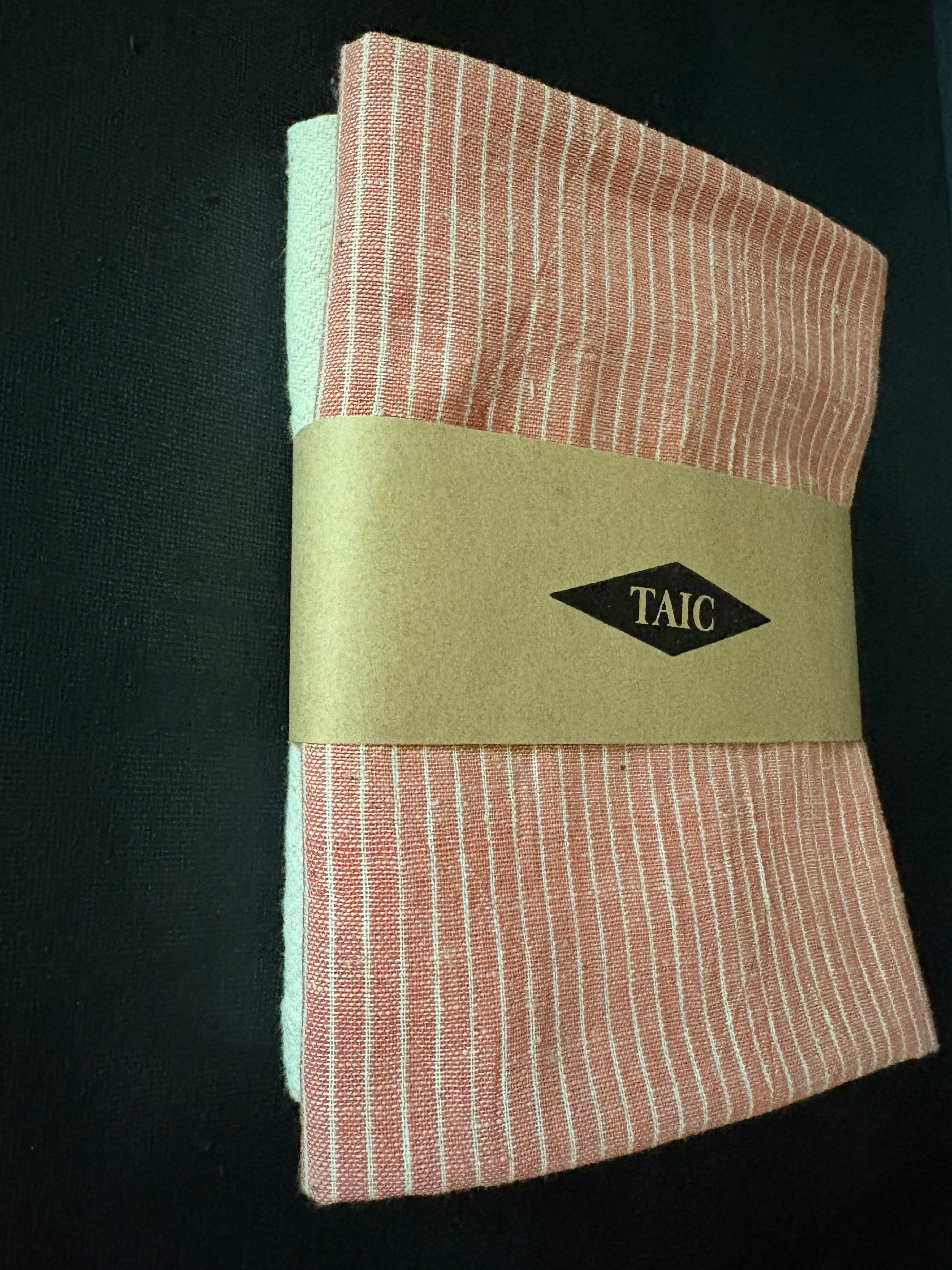 A folded cotton fabric set with thin vertical pink and white stripes, wrapped in a tan paper band featuring a black diamond logo with the text 'TAIC'.