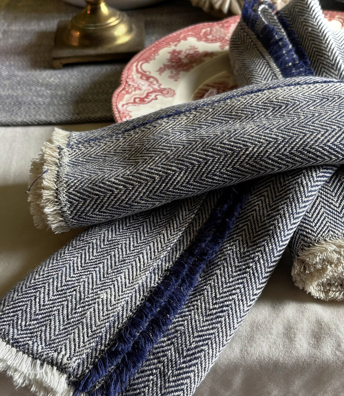 A close-up of a folded handwoven kala cotton textile napkin with a herringbone pattern in blue and white, with a dark blue stripe running through the center and frayed white edges, styled on a light surface with a decorative plate and brass object in the background.