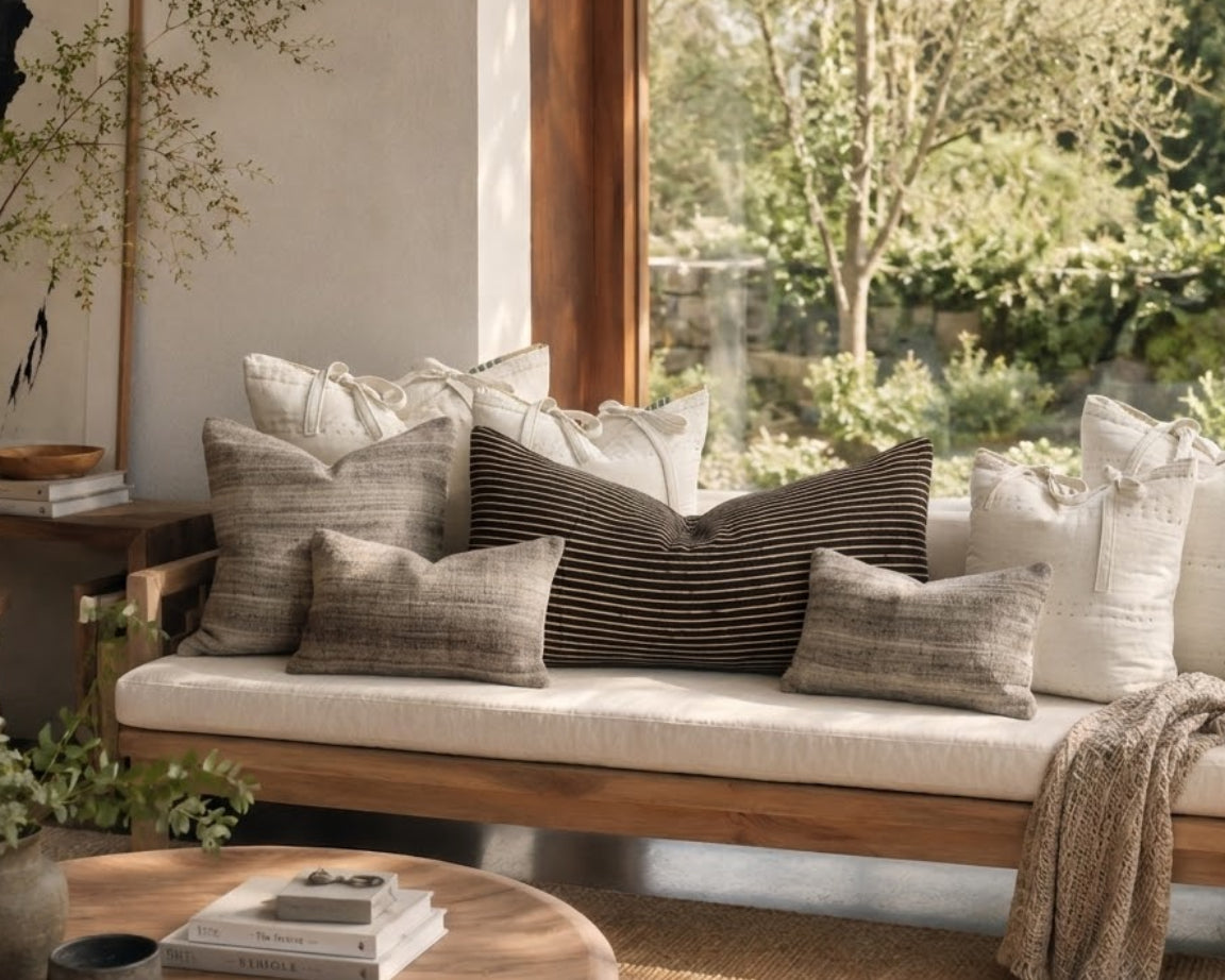 A wooden daybed with a cream upholstered cushion and multiple decorative throw pillows, styled in a bright room with a large window showing a garden view. A round wooden coffee table with books is in the foreground, and a beige throw blanket is draped over the right side.