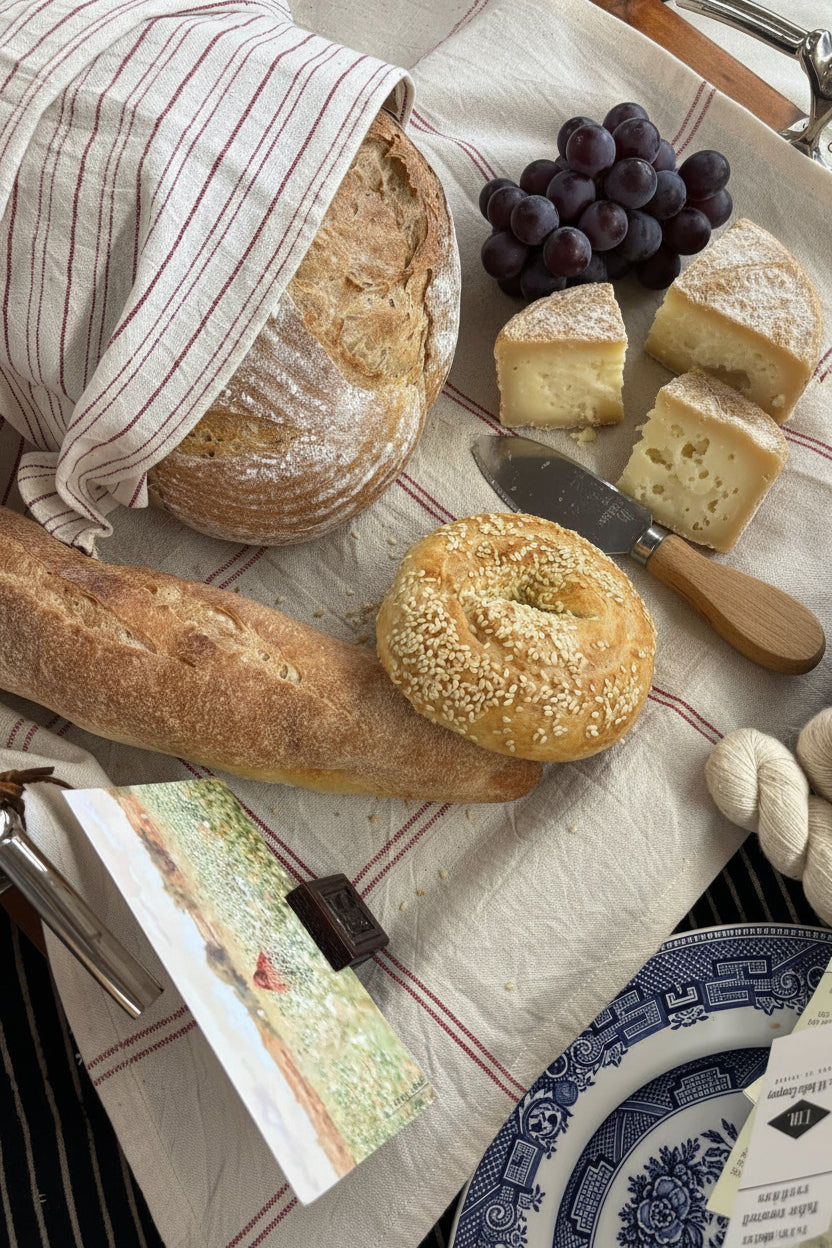 A styled picnic-style food arrangement on a striped Kala Cotton charcuterie liner, including a loaf of bread wrapped in a Kala Cotton bread basket liner, apples, and cheese.