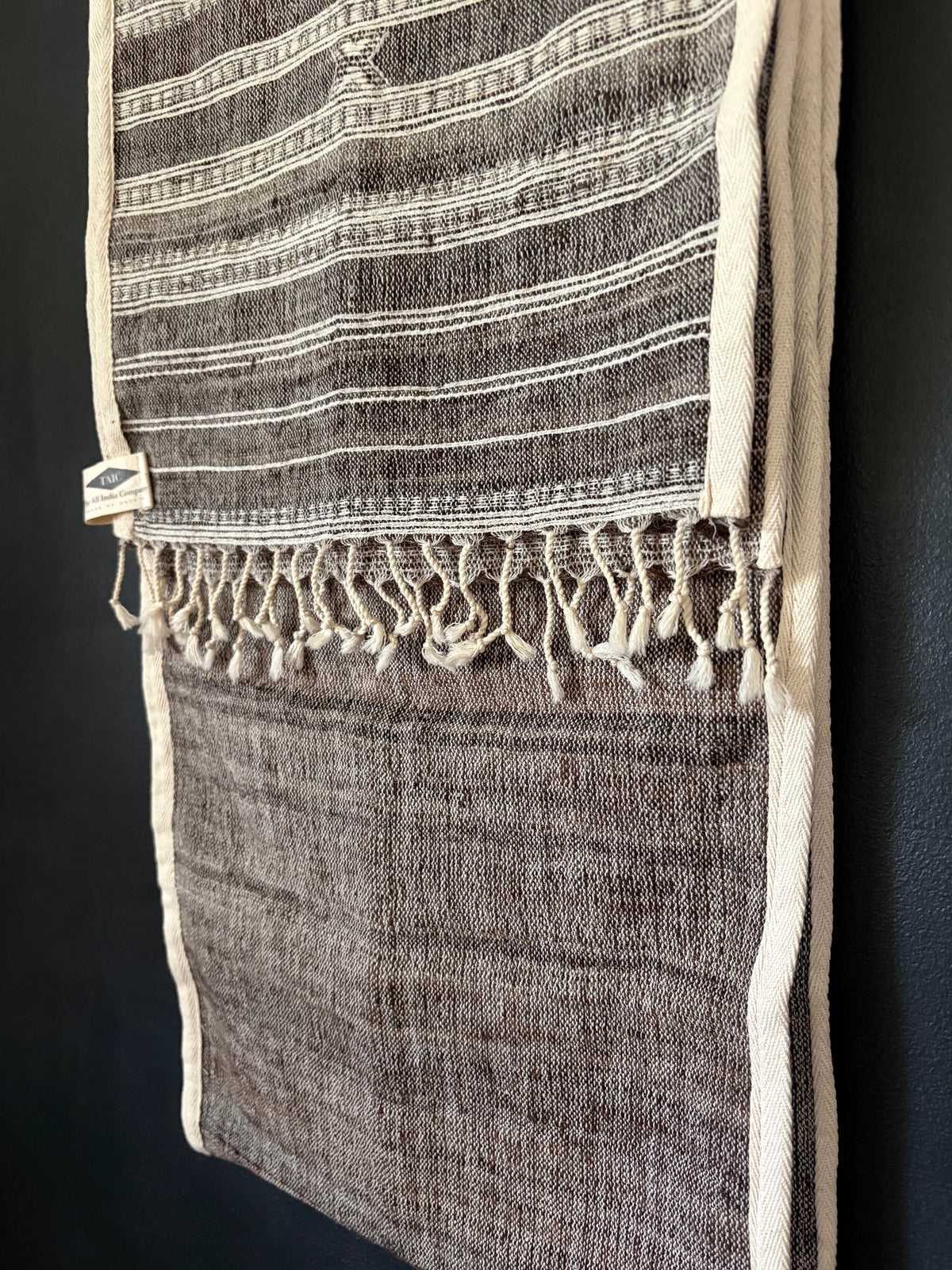 A dark brown and beige striped woven textile runner with cream-colored fringe and borders, hanging against a dark background.