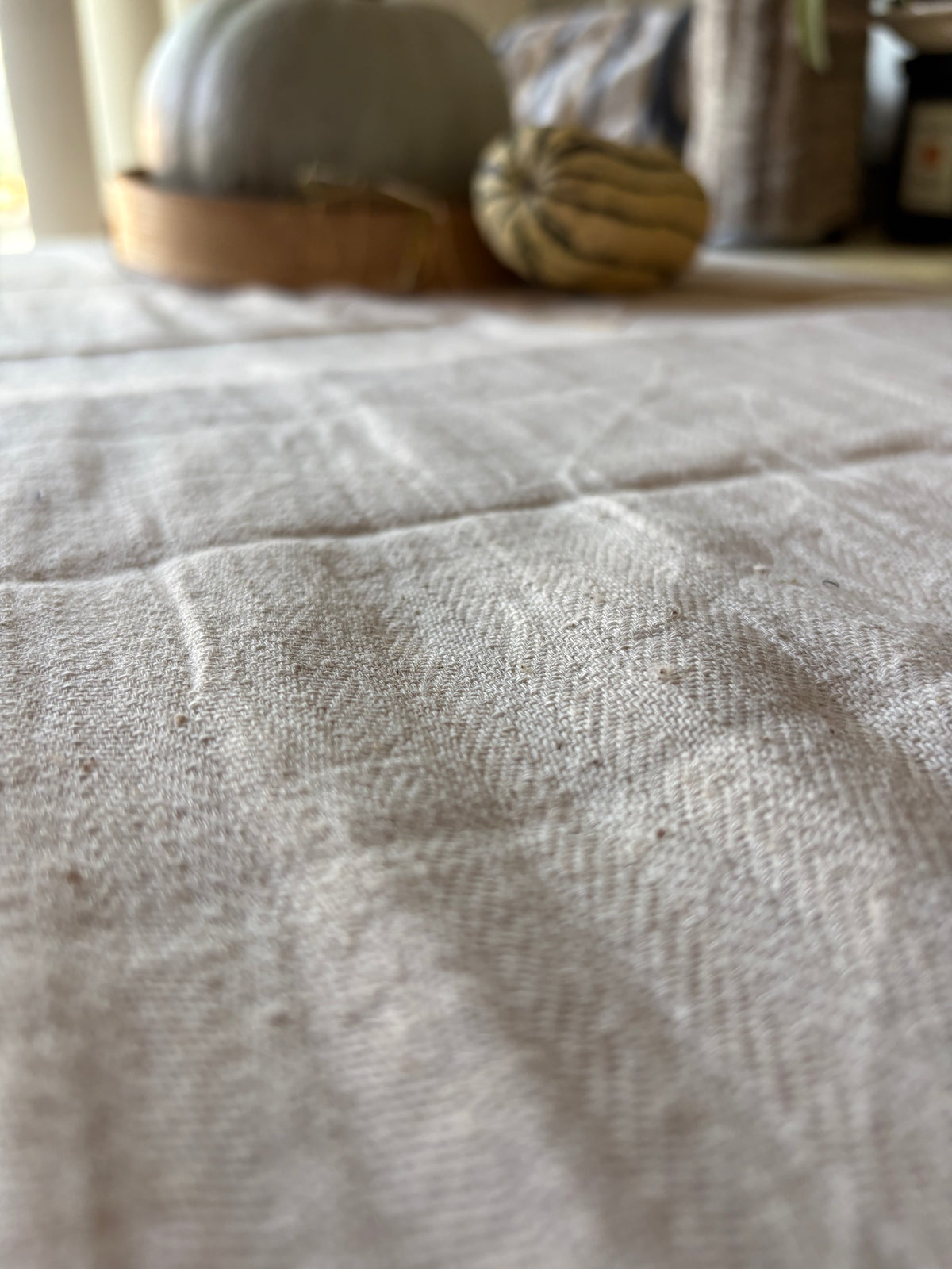 A close-up of a light beige linen tablecloth with a visible woven texture, with blurred decorative pumpkins in the background.