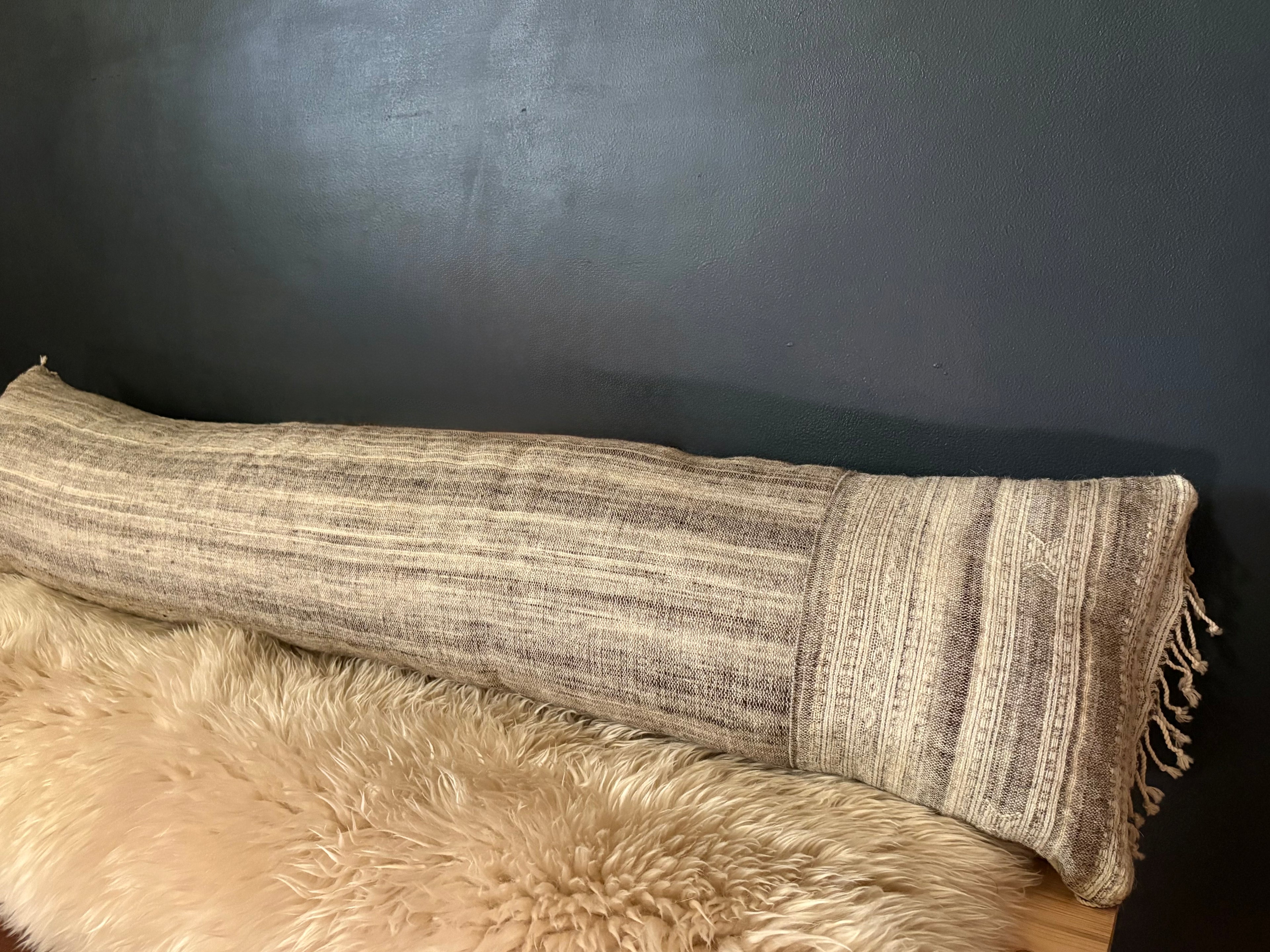 A long cylindrical lumbar pillow with a woven beige and brown striped pattern and fringe tassels, resting on a fluffy cream faux fur rug against a dark gray wall.