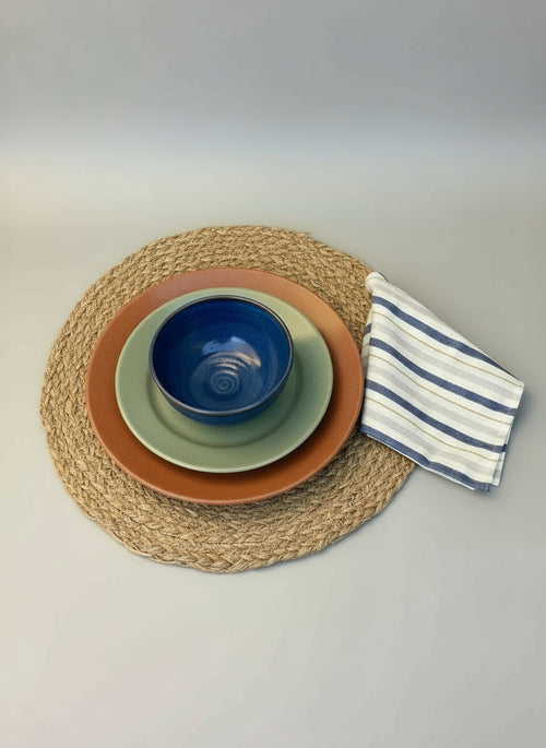 Set of green ceramic bowls on a round placemat with a striped cloth napkin on a light gray background